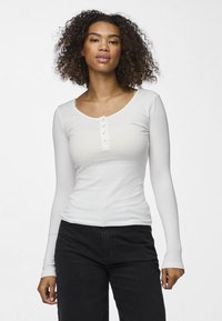 Bílý žebrovaný top s dlouhými rukávy, kulatým výstřihem a zapínáním na knoflíky. Má přiléhavý střih a jemnou texturu. S kombinací s černými kalhotami.