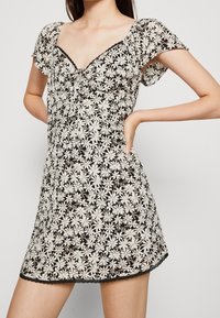 Robe courte à fleurs avec un fond noir et des motifs de marguerites blanches. Elle a des manches courtes, un décolleté en V et un ourlet avec de la dentelle.
