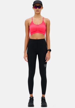 Frau trägt einen pinkfarbenen Sport-BH, schwarze hoch taillierte Leggings mit Logo, schwarze Sneaker, schwarze Kappe, Sonnenbrille und eine Smartwatch, steht aufrecht.