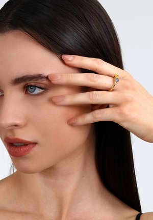 Goldring mit einem blauen Stein, verfügt über ein elegantes Band und aufwendige Details. Die Finger des Modells zeigen einen natürlichen Nagellack.