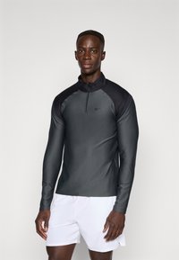 T-shirt de sport à manches longues en gris foncé, avec un col zippé à quart, un tissu texturé et une coupe élégante. Associé à un short blanc.