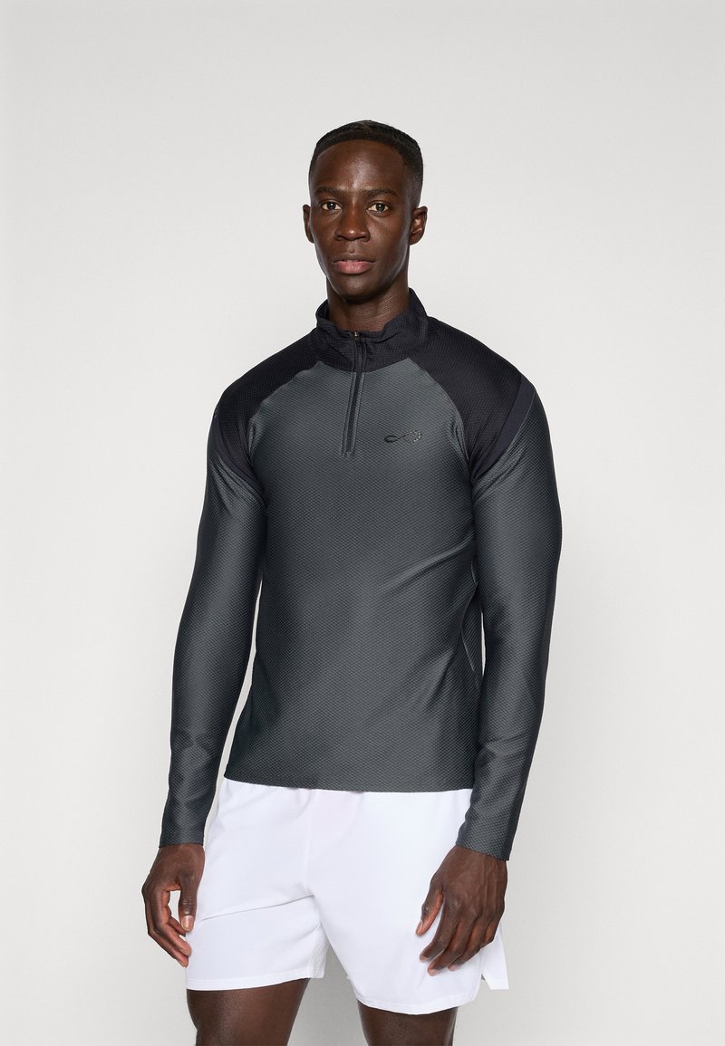 T-shirt de sport à manches longues en gris foncé, avec un col zippé à quart, un tissu texturé et une coupe élégante. Associé à un short blanc.