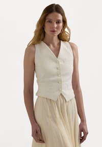 Colete de linho branco com um decote em V profundo, quatro botões e dois bolsos frontais, combinado com uma saia leve e texturizada. Tecido suave e natural.