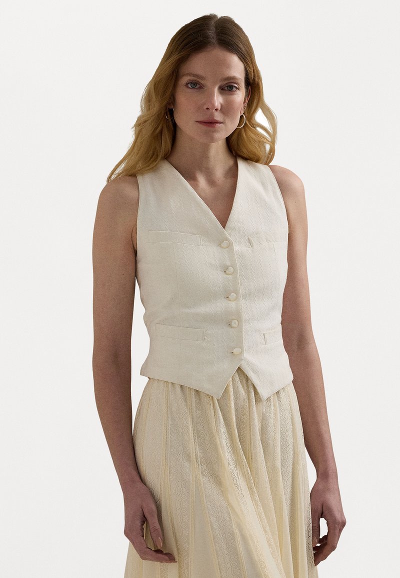 Colete de linho branco com um decote em V profundo, quatro botões e dois bolsos frontais, combinado com uma saia leve e texturizada. Tecido suave e natural.