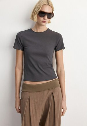 Femeie cu păr scurt și blond, purtând ochelari de soare mari și întunecați, bluză mulată cu mâneci scurte de culoare gri închis și pantaloni largi maro cu talie lată.