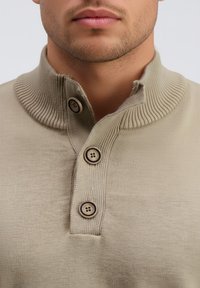 Suéter beige con cuello con ribete y tres botones marrones. El material parece suave con una textura lisa. Vista en primer plano.