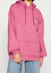 Rosa huvtröja i mjukt tyg med en kenguruficka, löst snitt och dragsko-accenter. Har en broderad logotyp på bröstet.