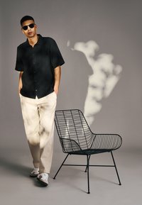 Camicetta nera a maniche corte, pantaloni beige e occhiali da sole indossati da una persona affianco a una sedia in filo nero dal design moderno su uno sfondo neutro.