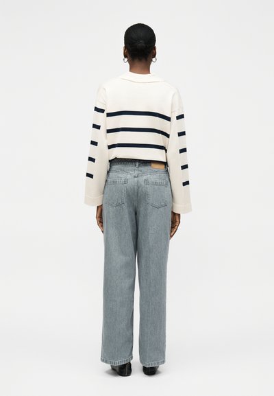 Άτομο με τα μαλλιά σε κότσο, φοράει λευκό πουλόβερ με μαύρες οριζόντιες ρίγες και φαρδιά ανοιχτό μπλε τζιν, εμφανιζόμενο από πίσω σε λευκό φόντο.
