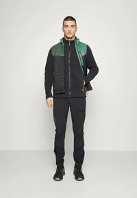 Male model in black zip-up jacket, black pants, green and black sleeveless vest, and black shoes standing against plain background.