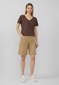 T-shirt marron à col en V associé à un short en khaki retroussé. Le modèle porte des sneakers de couleur claire. Le tissu semble doux avec une coupe décontractée.