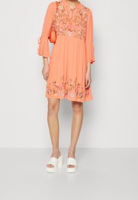 Korallenfarbenes Kleid mit floraler Stickerei, Bindekragen und Glockenärmel. Es hat einen ausgestellten Saum und wird mit weißen Plateausandalen kombiniert.