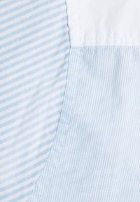 Tessuto azzurro chiaro con sottili righe bianche, che presenta una texture liscia. Il motivo si trasforma in tessuto blu uniforme sul bordo, mostrando una cucitura pulita.