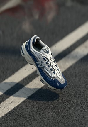 Białe i niebieskie buty sportowe z siateczkową cholewką, wyściółką w kołnierzu i widoczną amortyzacją. Posiadają elementy odblaskowe oraz teksturowaną podeszwę.