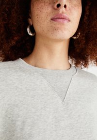 sweatshirt gris avec une finition texturée, arborant un décolleté en V et un léger détail de couture, porté avec une boucle d'oreille en argent.