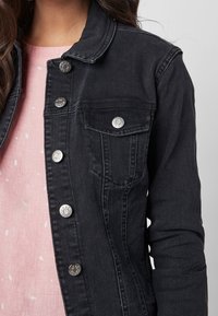 Black denim jacket featuring a buttoned front, chest pocket, and silver hardware accents. Background shows a light pink top with white patterns.