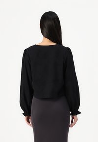 Blouse noire courte avec manches longues bouffantes et un col rond, fabriquée en tissu texturé. Portée avec une jupe grise ajustée.