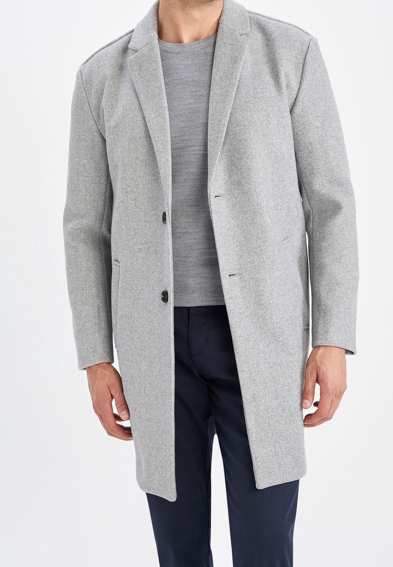 Grå uldfrakke med spids krave, enkelt knaplukning og to forlommer. Båret sammen med en grå tekstureret sweater nedenunder.
