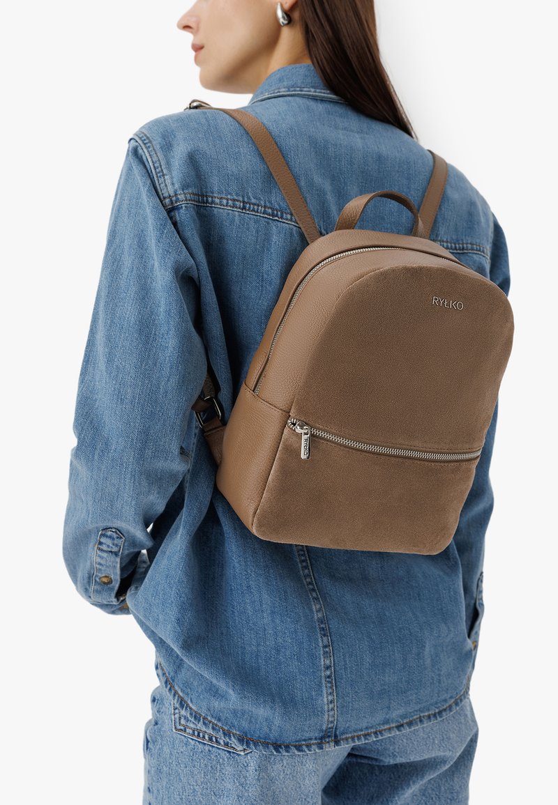 Bruine neplederen rugzak met een afgeronde vorm, voorzak met rits en verstelbare banden, gedragen over een spijkerjack.