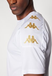 Camisa atlética blanca con tejido texturizado y gráficos en dorado en el hombro. Diseño con escote suave y mangas cortas.