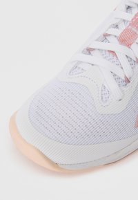 Nahaufnahme eines weißen Sportschuhs mit Mesh-Stoff, weißen Schnürsenkeln, pinken Akzenten und beigem Sohlen auf einem einfarbigen hellen Hintergrund.