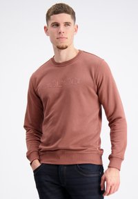 Sudadera de algodón marrón con mangas largas, cuello y puños de canalé, con texto de logo en relieve en el pecho. Combinada con jeans azul oscuro.