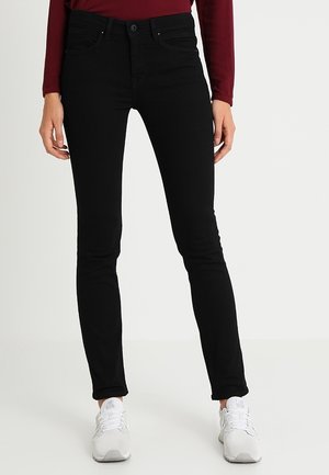 Jean noir slim fit porté avec un top bordeaux à manches longues et des chaussures de sport blanches, présenté de la taille aux pieds sur un fond uni.