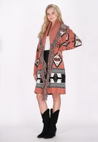 Gebreide cardigan in roestkleur met zwart-witte geometrische patronen. Heeft een brede kraag en franjes aan de voorkant.