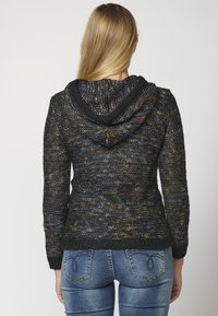 Žena s dlouhými blond vlasy, která má na sobě vícebarevnou pletenou mikinu s kapucí a modré džíny, stojící čelem k jednobarevnému bílému pozadí.