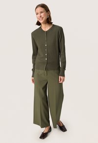 Olijfgroene geribde cardigan met een ronde halslijn, knoopsluiting en lange mouwen, gecombineerd met wijde olijfgroene broeken en bruine schoenen.