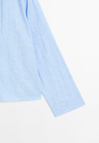 Chemise à manches longues bleu clair en tissu texturé. Présente une coupe décontractée et un design simple avec de légères pinces le long des manches.