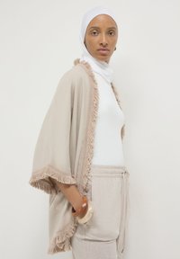 Donna che indossa un hijab bianco, una maglietta bianca, uno scialle beige con frange e pantaloni beige con coulisse, in posa con le mani nelle tasche su uno sfondo neutro.
