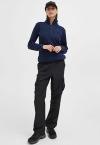 Pull en polaire bleu marine avec un col zippé mi-haut associé à un pantalon cargo noir ; comporte plusieurs poches et un tissu texturé. Des chaussures noires complètent le look.