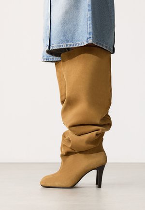 Tan suede slouchy knee-high boots with pointed toes and black stiletto heels worn with light blue denim jeans.