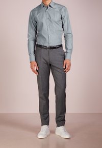 Camicia grigia con bottoni e maniche lunghe, abbinata a pantaloni eleganti grigio scuro e sneakers bianche. La camicia presenta un colletto classico e una texture liscia.