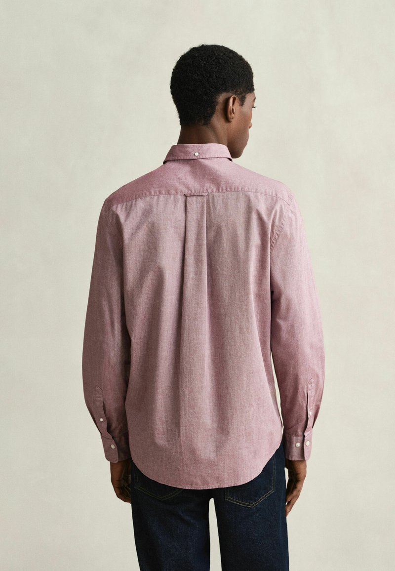 Homme portant une chemise rose à manches longues et un jean foncé, vu de dos sur un fond uni clair.
