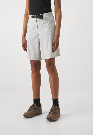 Person iført lysegrå knælange shorts med sort bælte, sort top, sorte sokker og brune vandresko står på ensfarvet baggrund.