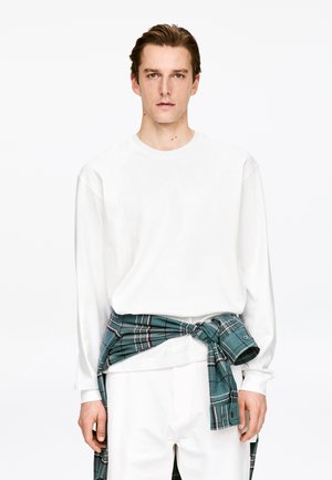 Giovane uomo che indossa una camicia bianca a maniche lunghe e pantaloni bianchi con una camicia a quadri verde annodata in vita, su uno sfondo bianco.