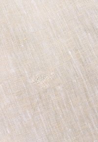 Lichtbeige linnenstof met een gestructureerde weving, waarbij subtiele onregelmatigheden en een delicate geborduurde detail in één gebied zijn te zien.