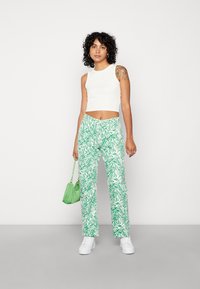 Top cropped branco combinado com umas calças largas verdes com um padrão de ondas. A modelo segura uma pequena mala verde e usa ténis brancos.
