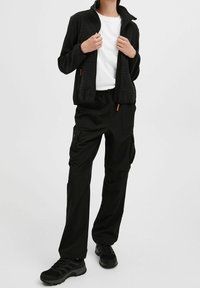 Czarna kurtka zapinana na zamek o teksturowanej powierzchni, w zestawieniu z czarnymi spodniami cargo z wieloma kieszeniami. Noszona z czarnymi butami sportowymi.