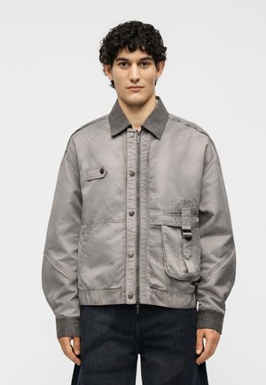 Άνδρας με σγουρά μαλλιά που φορά γκρι ζακέτα με φερμουάρ, γιακά με κουμπιά και καπάκι τσέπης, στέκεται μπροστά σε απλό φόντο.