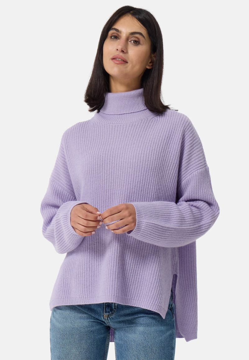 Pull en tricot lilas avec un col haut et une texture côtelée. Dispose de manches larges et de fentes sur les côtés, associé à un jean en denim bleu.