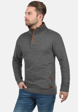 Grijze sweatshirt met een hoge kraag, voorzien van bruine accenten en knopen, een voorkeurszak en geribde manchetten. Draagt met blauwe jeans.