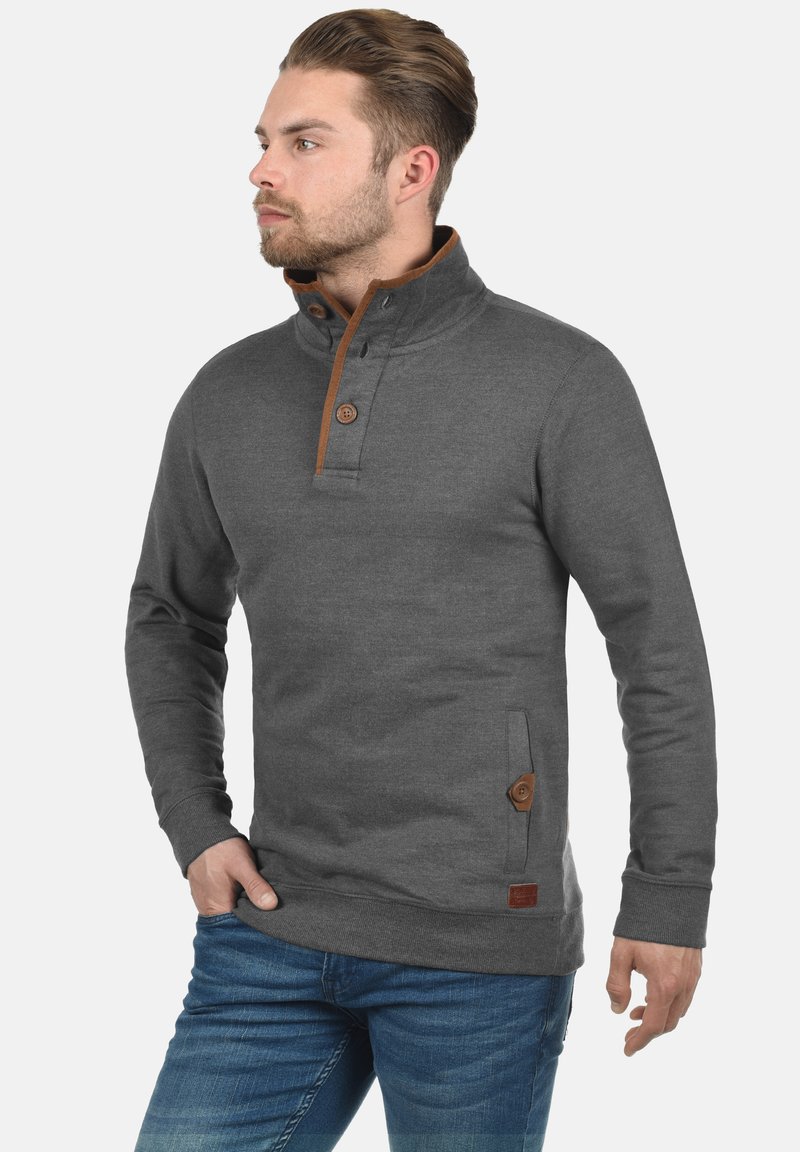 Sweatshirt gris avec un col montant, présentant des accents et des boutons marron, une poche avant et des poignets côtelés. Porté avec un jean bleu.