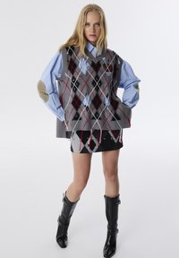 Grauer gestrickter Pullover mit Rautenmuster, roten und schwarzen Akzenten, über einem hellblauen Shirt getragen, kombiniert mit einem schwarzen Rock und hohen schwarzen Stiefeln.