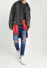Dżinsowa kurtka w wyblakłym czarnym kolorze, noszona na granatowej koszuli. Slim-fit niebieskie dżinsy z przetarciami na kolanach. Białe sneakersy dopełniają stylizację.
