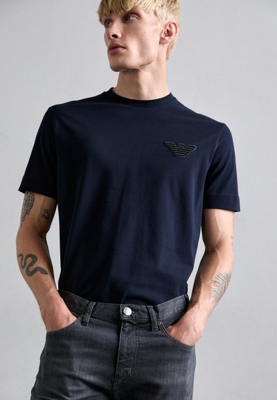 T-shirt girocollo blu navy in cotone, con un logo nero testurizzato sul petto. Abbinato a jeans in denim scuri.
