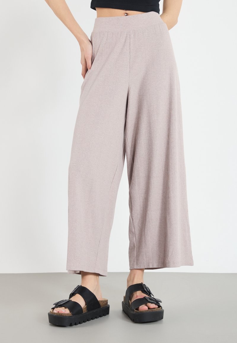 Personne portant un pantalon ample beige clair à jambes larges et des sandales noires à semelles épaisses, debout à l'intérieur devant un fond uni.