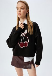 Zwarte gebreide trui met een kers en strik ontwerp in rood en wit. Draagt een witte blouse met kraag en een bordeauxleren rok.
